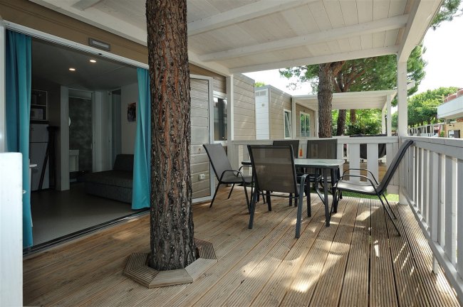 Un'ampia veranda in legno con tavolo e sedie, circondata da alberi.