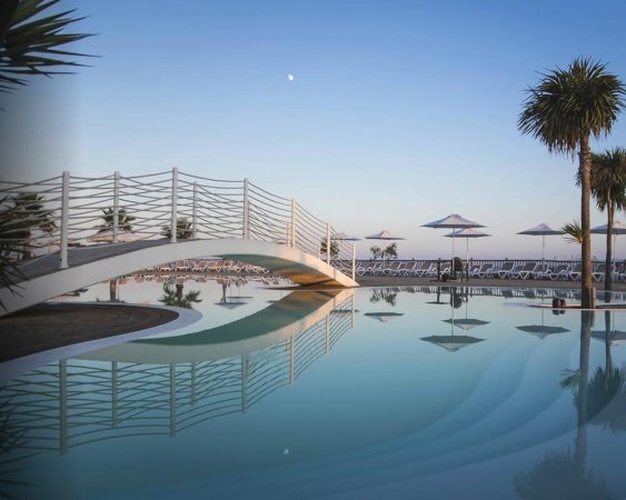 Piscina con ponte curvo e ombrelloni, alberi al tramonto.