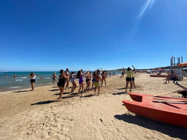 Persone che ballano sulla spiaggia in un giorno soleggiato. Il mare è visibile a sinistra.
