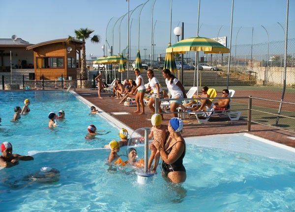 Persone in piscina con docce, alcune sedute su sdraio sotto ombrelloni.
