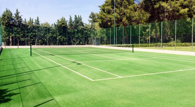 Campo da tennis all'aperto con erba sintetica verde e rete, circondato da alberi e recinzione.