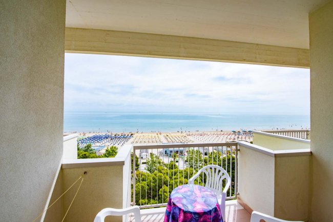 Un balcone con due sedie bianche e vista sul mare e sulla spiaggia con ombrelloni.