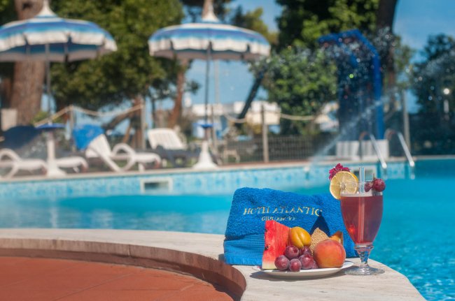 Bordo piscina con piatto di frutta, bevanda e asciugamano blu. Ombrelloni sullo sfondo.