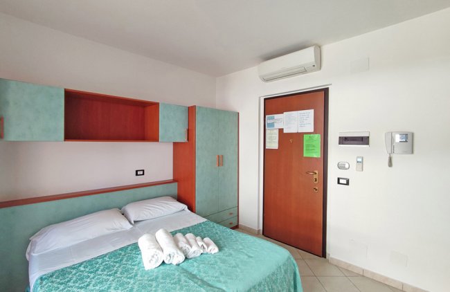 Letto matrimoniale con copriletto azzurro, armadio e porta d'ingresso con climatizzatore sopra.