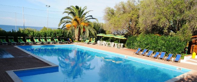 Piscina esterna con sedie a sdraio e ombrelloni, circondata da palme e vegetazione.