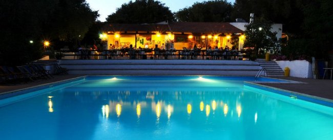 Piscina illuminata di notte con ristorante sullo sfondo.