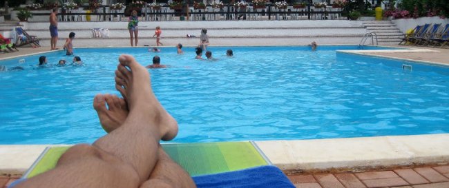 Gambe su lettino in piscina con persone che nuotano.