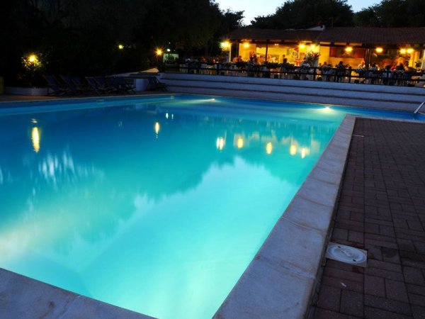 Piscina illuminata di sera con ristorante sullo sfondo.