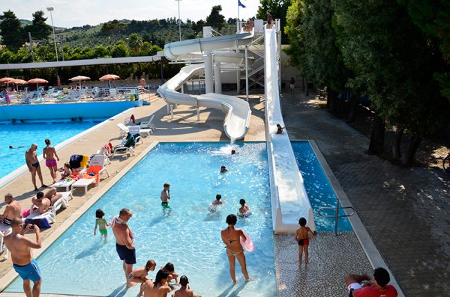 Piscina con scivoli d'acqua, persone che nuotano e si rilassano al sole.