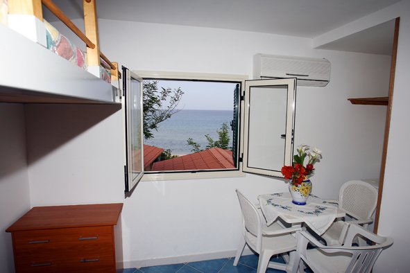 Interno di una stanza con tavolo e sedie. La finestra aperta offre vista sul mare.