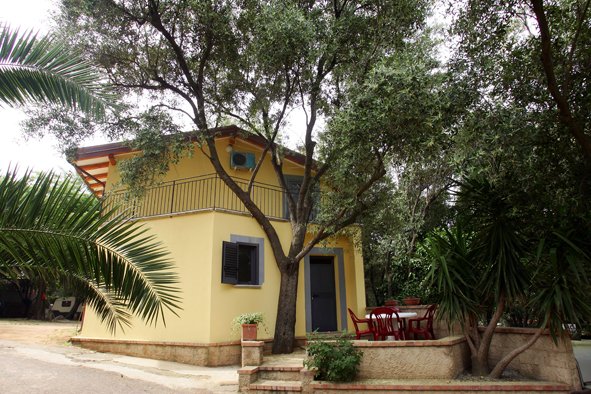Casa gialla con balcone, circondata da alberi e piante, con area seduta esterna.