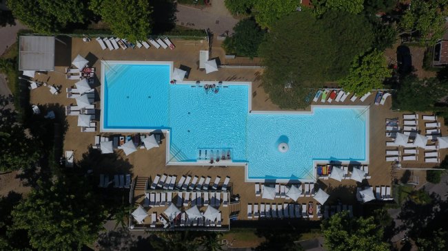 Piscina rettangolare azzurra vista dall'alto, circondata da lettini bianchi sotto ombrelloni.