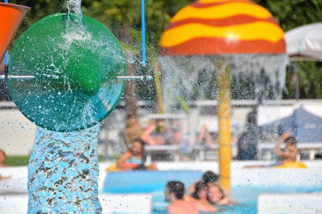 Un recipiente verde versa acqua in piscina, mentre persone nuotano sullo sfondo.