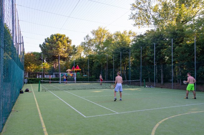 Due persone giocano a tennis su un campo all'aperto, circondato da alberi e una rete di protezione.