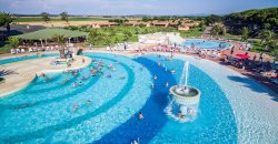 Ampia piscina a onde con fontana centrale, persone in acqua, circondata da vegetazione e strutture.