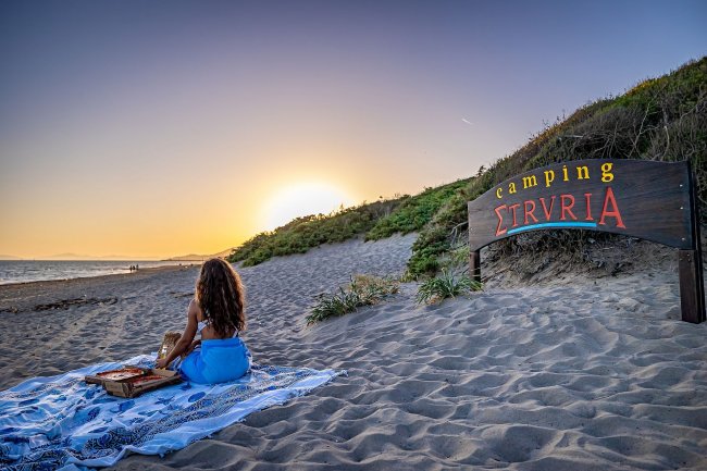 Tramonto su spiaggia con donna e cartello camping.
