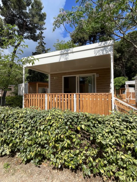 Un bungalow moderno con veranda e ringhiera in legno circondato da alberi e siepi verdi.