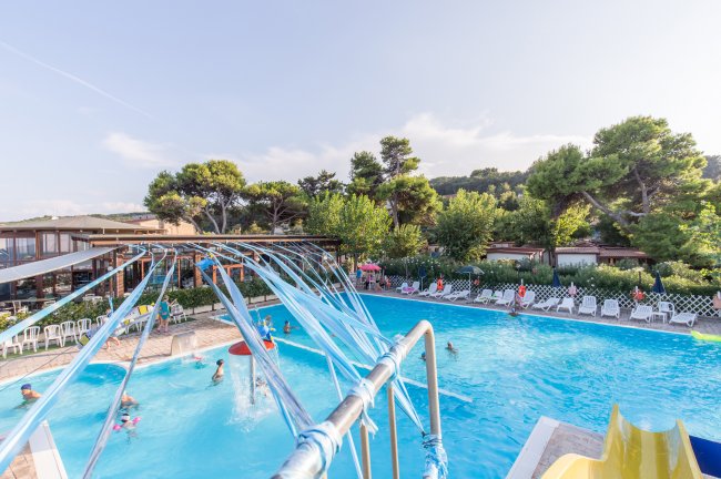 Piscina all'aperto con giochi d'acqua e persone che nuotano, circondata da alberi e sedie a sdraio.