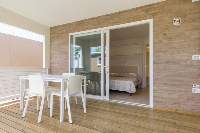 Terrazza con tavolo bianco e sedie, vista su camera da letto attraverso porta scorrevole.