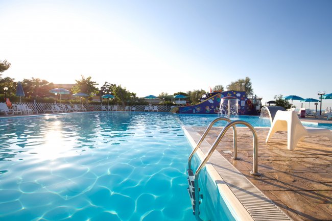 Piscina con scivolo colorato, ombrelloni e riflessi del sole sull'acqua.