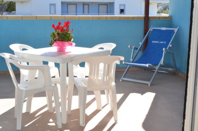 Tavolo bianco con sedie e pianta rossa su terrazza, sedie a sdraio blu sullo sfondo.