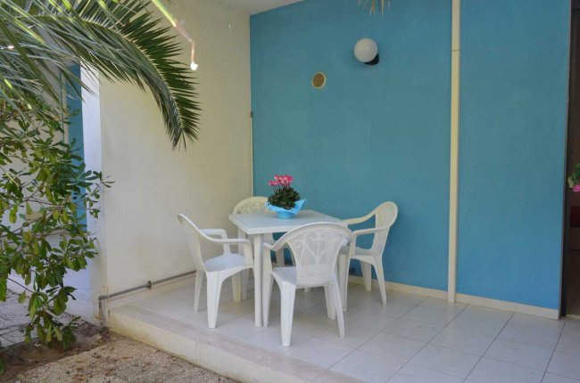 Tavolo bianco esterno con quattro sedie di plastica, parete blu e pianta verde.