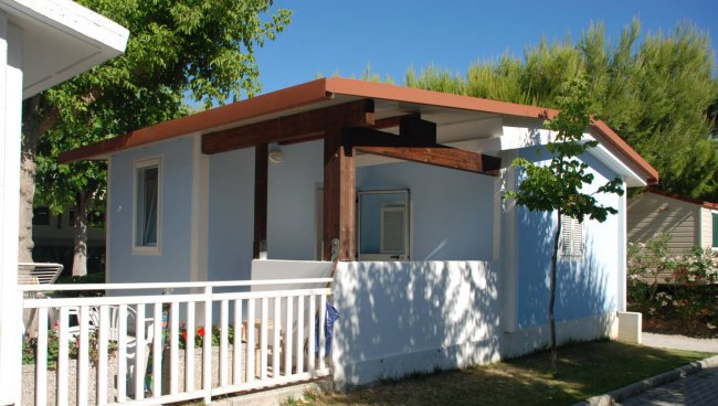 Bungalow bianco e azzurro con tetto rosso-marrone e un piccolo patio, circondato da alberi.