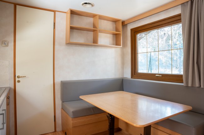 Area pranzo con tavolo in legno e sedute a L grigie, finestra e mensole vuote.