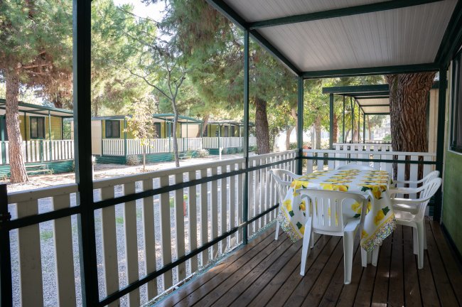 Veranda di legno coperta con tavolo e sedie bianche, circondata da alberi.