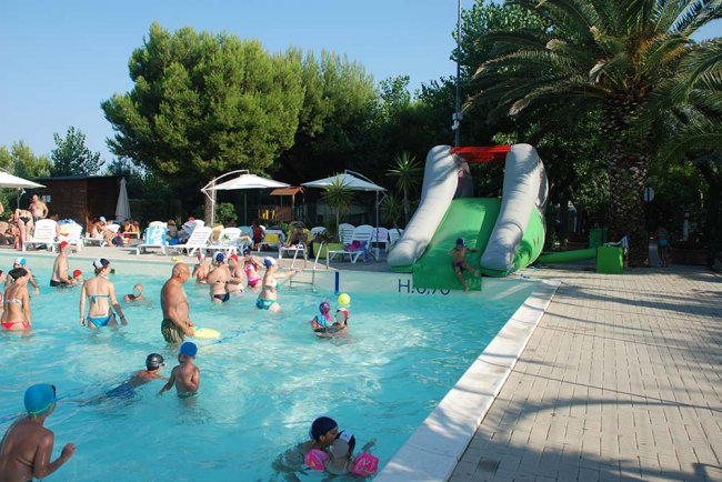 Una piscina gremita di persone con uno scivolo gonfiabile verde e sedie sdraio sotto ombrelloni.