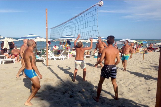 Persone giocano a pallavolo su una spiaggia soleggiata con ombrelloni e mare sullo sfondo.