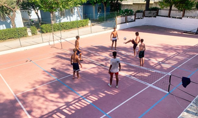 Persone giocano a footvolley su un campo con rete, circondato da alberi.