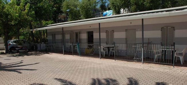 Una fila di bungalow all'ombra degli alberi, con sedie bianche davanti.