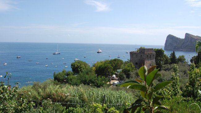 Costa con mare blu, vegetazione, barche e una torre antica in pietra.