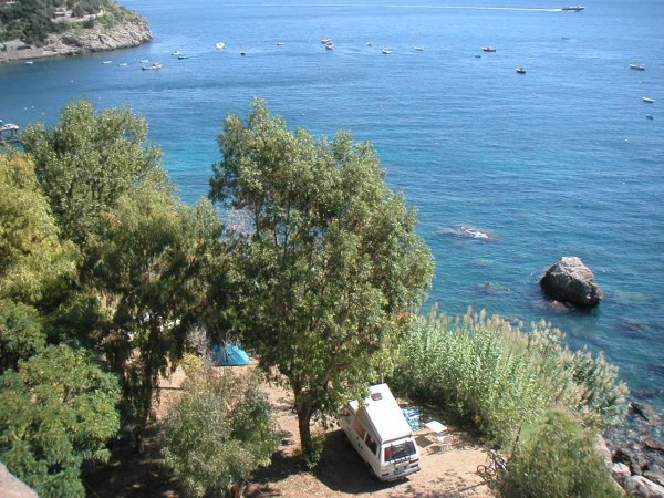 Vista dall'alto di un campeggio su una costa rocciosa con alberi e mare.
