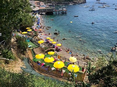 Spiaggia con ombrelloni gialli, mare azzurro e barche al largo. Persone prendono il sole e nuotano.