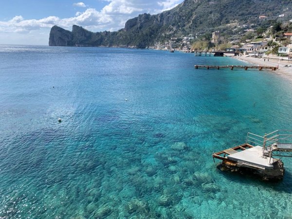Costa con acque turchesi, un molo e colline verdi in lontananza.