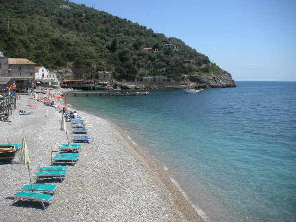 Spiaggia di sassi con lettini blu, mare calmo e colline verdi sullo sfondo.