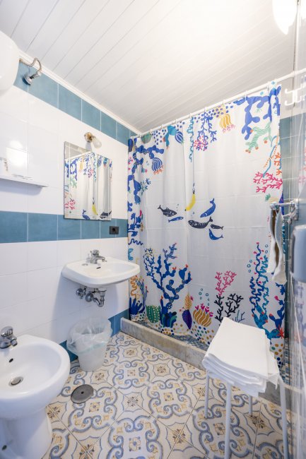 Bagno con piastrelle blu e bianche, doccia con tenda colorata a tema marino e lavabo con specchio.