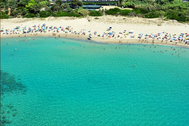 Spiaggia con ombrelloni colorati, bagnanti e mare verde chiaro. Vegetazione sullo sfondo.