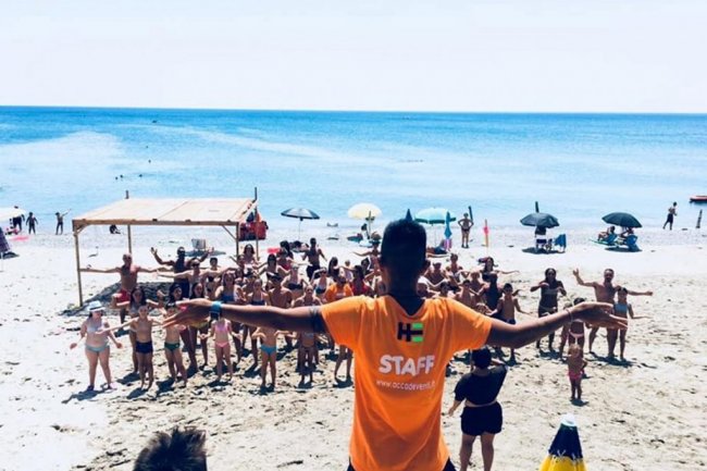 Persona in maglietta arancione guida un gruppo su una spiaggia.