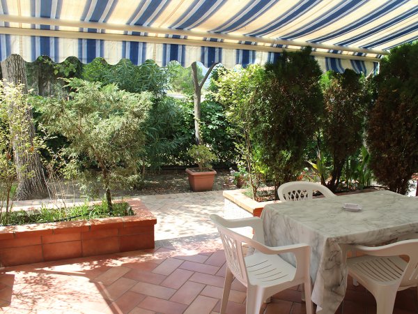 Un patio coperto con tavolo e sedie in plastica, circondato da piante e vasi su piastrelle.