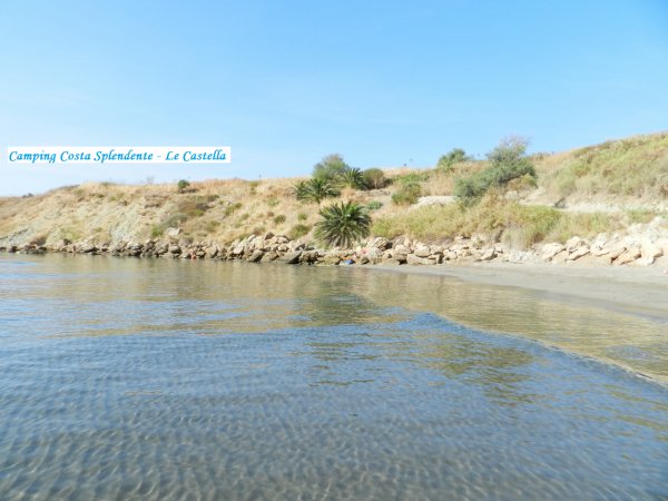Spiaggia con acqua calma, colline erbose e un cartello di campeggio in lontananza.