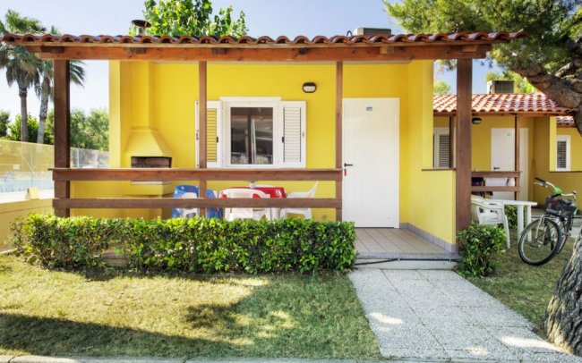 Una casa gialla con veranda in legno, sedie colorate e un barbecue nel giardino.