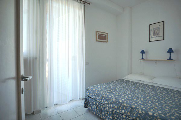 Letto matrimoniale con copriletto blu e giallo, mensola con lampade blu, finestra con tende bianche.