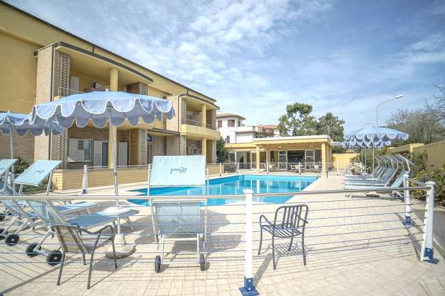 Piscina all'aperto con lettini e ombrelloni blu, edificio giallo sullo sfondo.
