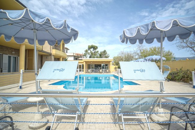 Vista frontale di una piscina con ombrelloni e lettini blu, cielo parzialmente nuvoloso.