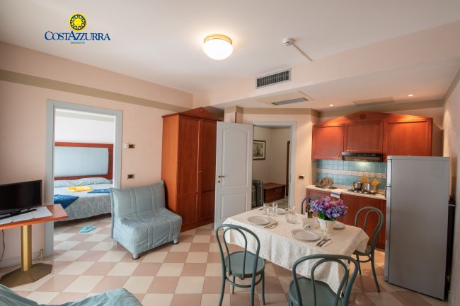Interno di un appartamento con soggiorno, cucina, tavolo apparecchiato e camera da letto.