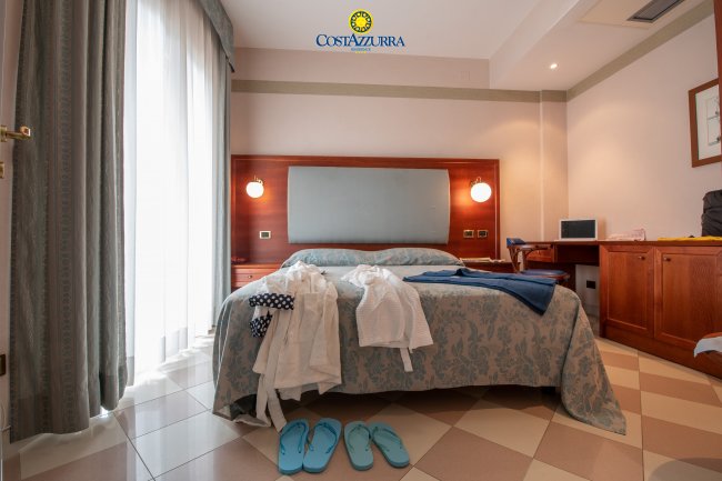 Camera d'albergo con letto matrimoniale, tende bianche, ciabatte azzurre e vestiti sparsi.