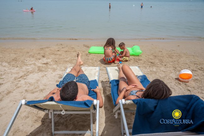 Spiaggia con persone su sdraio e bambini che giocano sulla sabbia.
